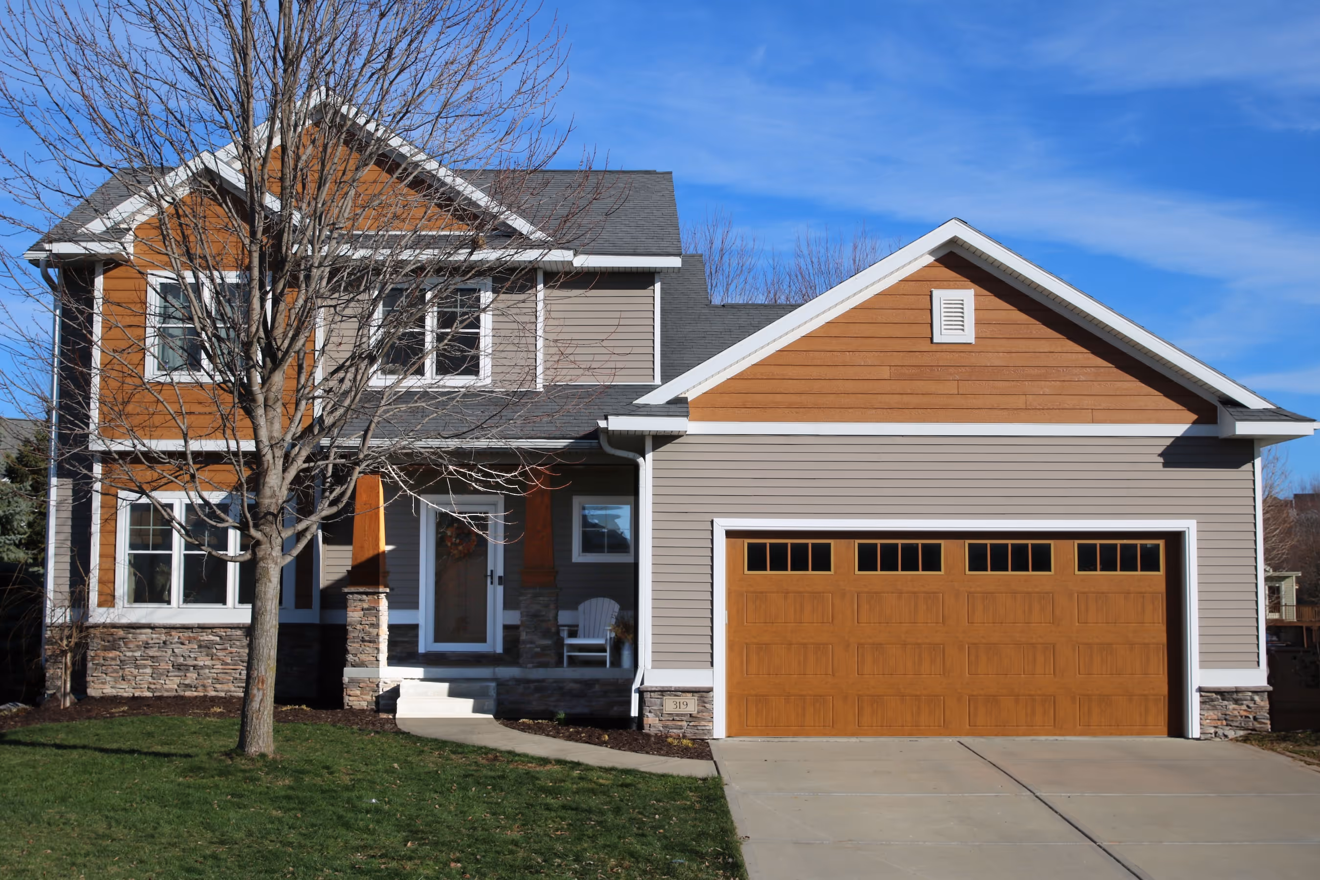 Two-story suburban house with gray and wood-toned siding, a large garage, a leafless tree in front, and clear blue sky.